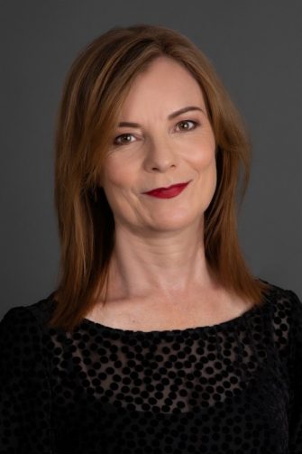 A photo of Felicity Plunkett, a white middle-aged woman. She has shoulder-length brown hair, and wears deep red lipstick. She is smiling.