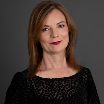 A photo of Felicity Plunkett, a white middle-aged woman. She has shoulder-length brown hair, and wears deep red lipstick. She is smiling.