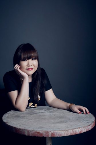 A photo of Julie Koh, an Australian Chinese-Malaysian woman. She sits at a stone table, her chin resting on her hand/