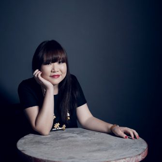 A photo of Julie Koh, an Australian Chinese-Malaysian woman. She sits at a stone table, her chin resting on her hand/