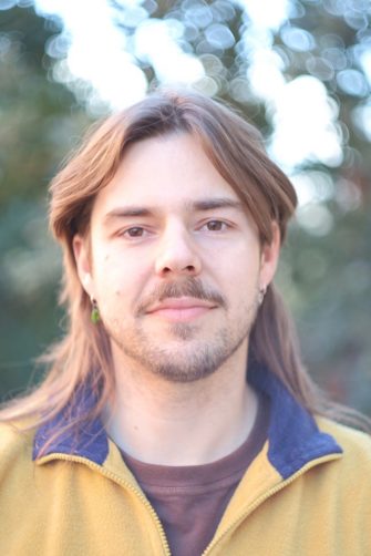 A photo of Troy Reid, a First Nations Dharug man. He is young, with shoulder-length brown hair and a light beard.