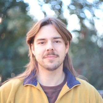 A photo of Troy Reid, a First Nations Dharug man. He is young, with shoulder-length brown hair and a light beard.