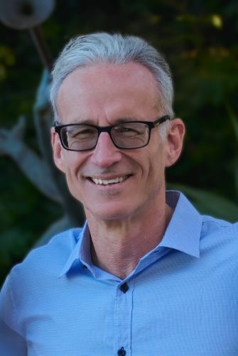 A photograph of academic Chris Conti. He is a middle-aged white man with short grey hair and glasses. He is smiling brightly, on a background of greenery.
