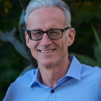 A photograph of academic Chris Conti. He is a middle-aged white man with short grey hair and glasses. He is smiling brightly, on a background of greenery.