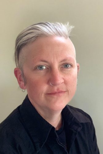 A photograph of author Jennifer Mills. She is a middle-aged white woman with short grey and black hair. She has a nose piercing and ear piercings.