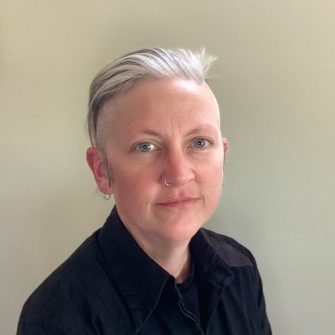 A photograph of author Jennifer Mills. She is a middle-aged white woman with short grey and black hair. She has a nose piercing and ear piercings.