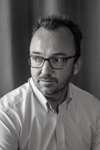 A photograph of Luke Johnson. He is a middle-aged white man with short, dark hair, and a short beard. He wears glasses. He is looking contemplatively to the side.