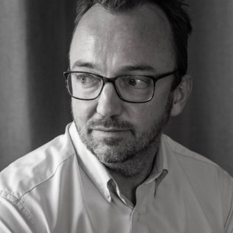 A photograph of Luke Johnson. He is a middle-aged white man with short, dark hair, and a short beard. He wears glasses. He is looking contemplatively to the side.