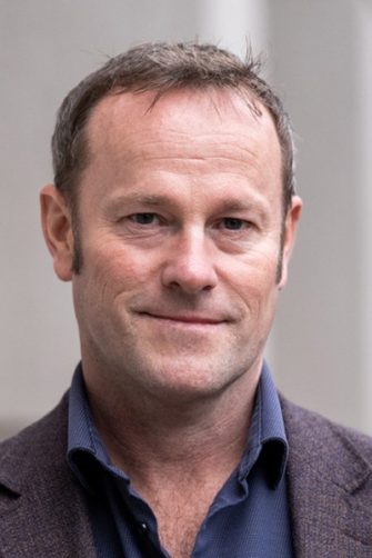 A photograph of academic Nathan Hollier. He is a middle-aged white man with short, dark hair. He is wearing a suit and smiling gently.