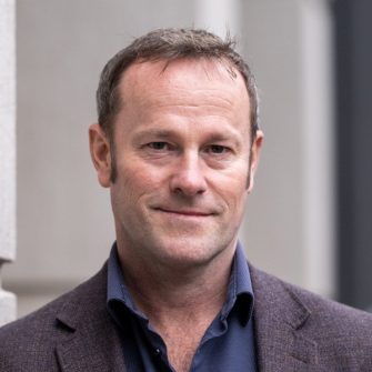 A photograph of academic Nathan Hollier. He is a middle-aged white man with short, dark hair. He is wearing a suit and smiling gently.