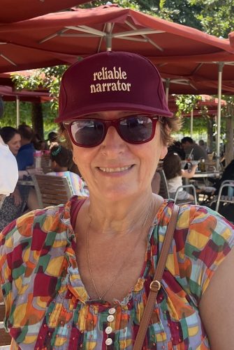 A photograph of Sue Joseph. She is a middle-aged white woman with short brown hair. She wears dark sunglasses, and a red cap which reads "reliable narrator". She smiles brightly.