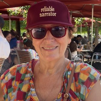 A photograph of Sue Joseph. She is a middle-aged white woman with short brown hair. She wears dark sunglasses, and a red cap which reads "reliable narrator". She smiles brightly.
