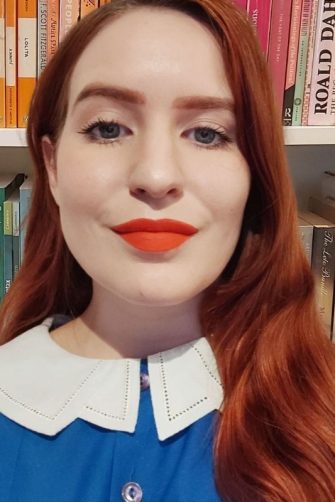 A photograph of Lizzie Levot. She is a young white woman with long red hair. She wears a blue shirt and bright red lipstick.