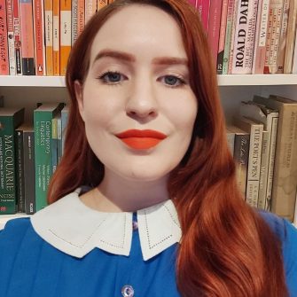 A photograph of Lizzie Levot. She is a young white woman with long red hair. She wears a blue shirt and bright red lipstick.