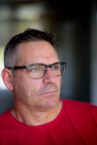 A photo of Lee McGowan. He is a middle-aged white man, with short black and grey hair. He wears glasses, and a bright red t-shirt. He looks contemplatively to the side.