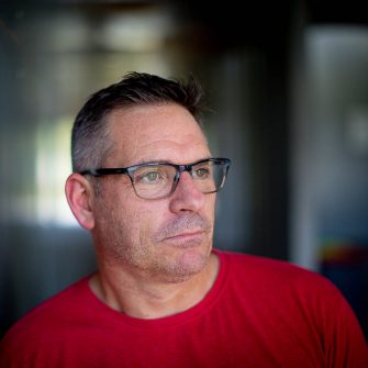 A photo of Lee McGowan. He is a middle-aged white man, with short black and grey hair. He wears glasses, and a bright red t-shirt. He looks contemplatively to the side.