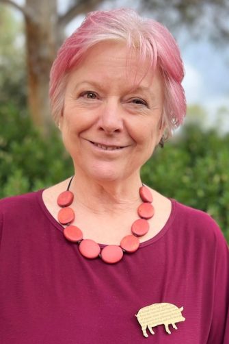 A photograph of Jen Webb. She is a middle-aged white woman. She has white hair, dyed pink. She wears a deep pink shirt and pink necklace.