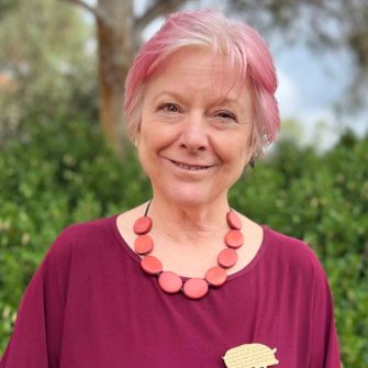 A photograph of Jen Webb. She is a middle-aged white woman. She has white hair, dyed pink. She wears a deep pink shirt and pink necklace.