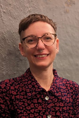 A photo of Ariella Van Luyn, a white nonbinary academic with short brown hair and glasses. They are smiling brightly.