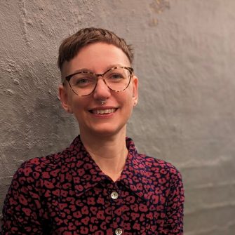 A photo of Ariella Van Luyn, a white nonbinary academic with short brown hair and glasses. They are smiling brightly.