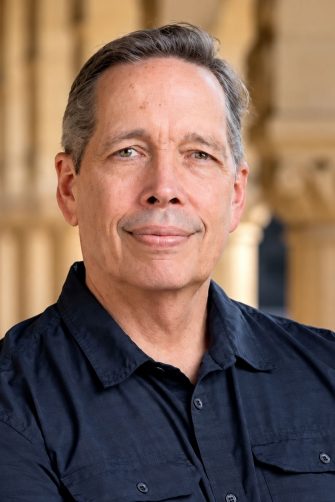 Professional headshot of a man wearing a dark navy shirt, with sandstone columns softly blurred in the background