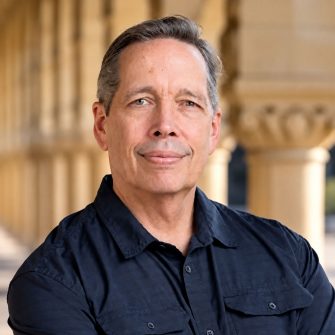 Professional headshot of a man wearing a dark navy shirt, with sandstone columns softly blurred in the background